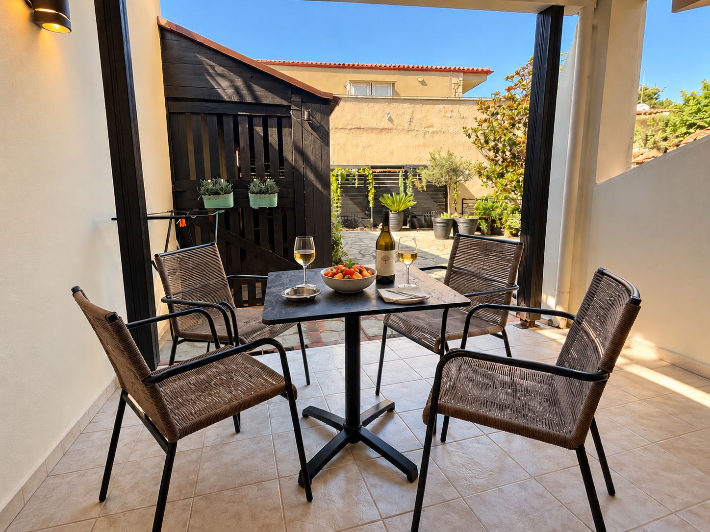 Een terras buiten met een tafel gedekt voor twee personen, omringd door rieten stoelen, een fles wijn en een schaal fruit.