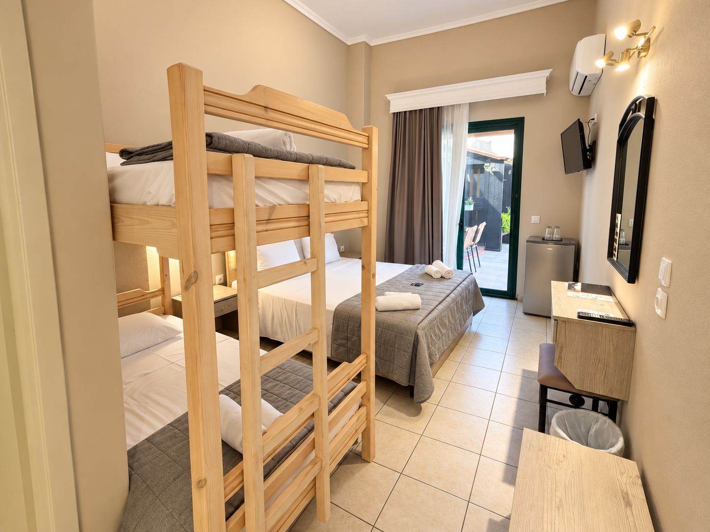 A hotel room with bunk beds, a desk, a mirror, and a view of the outside.