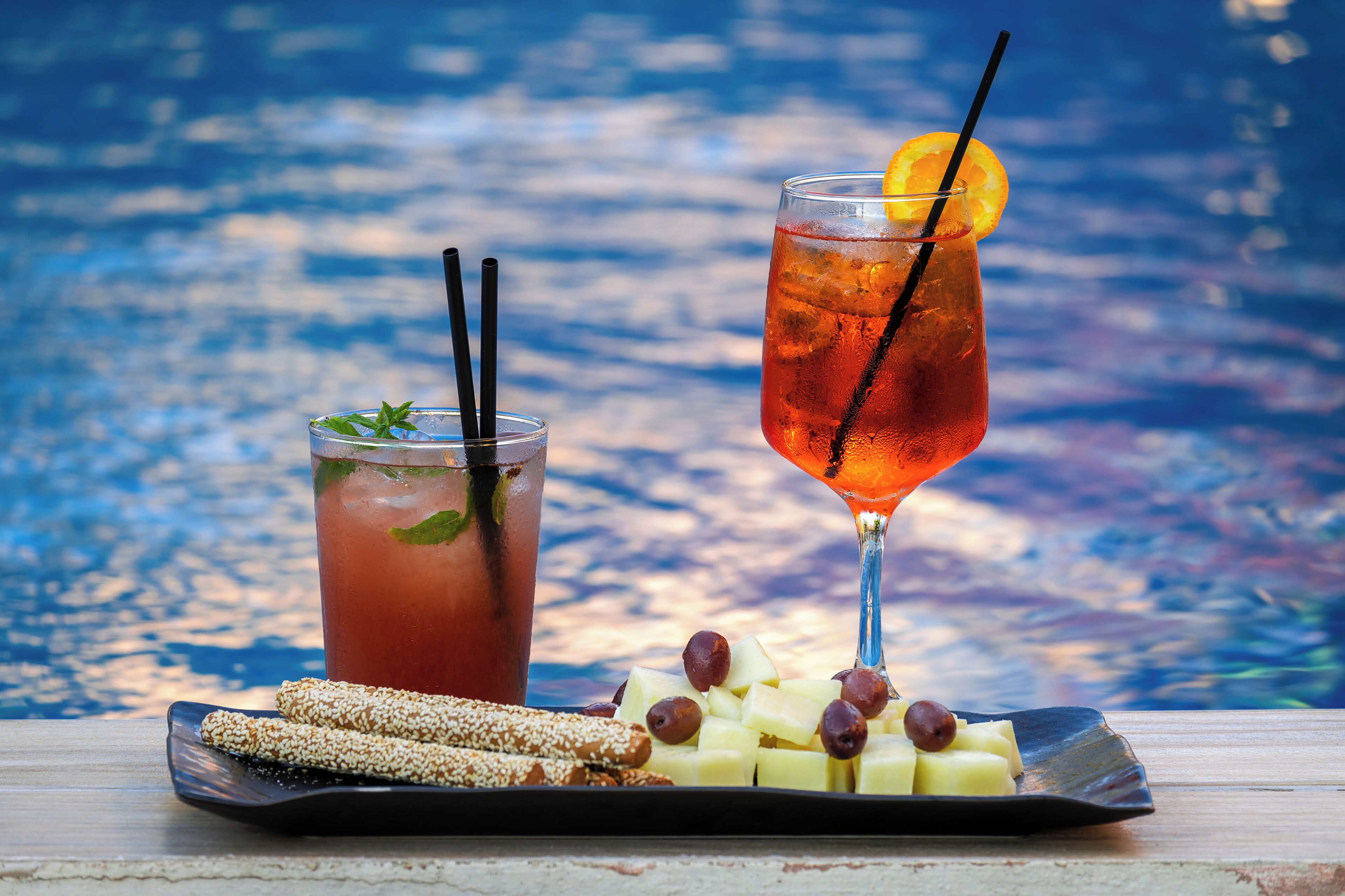 Two drinks with garnishes and a plate of snacks by a poolside.