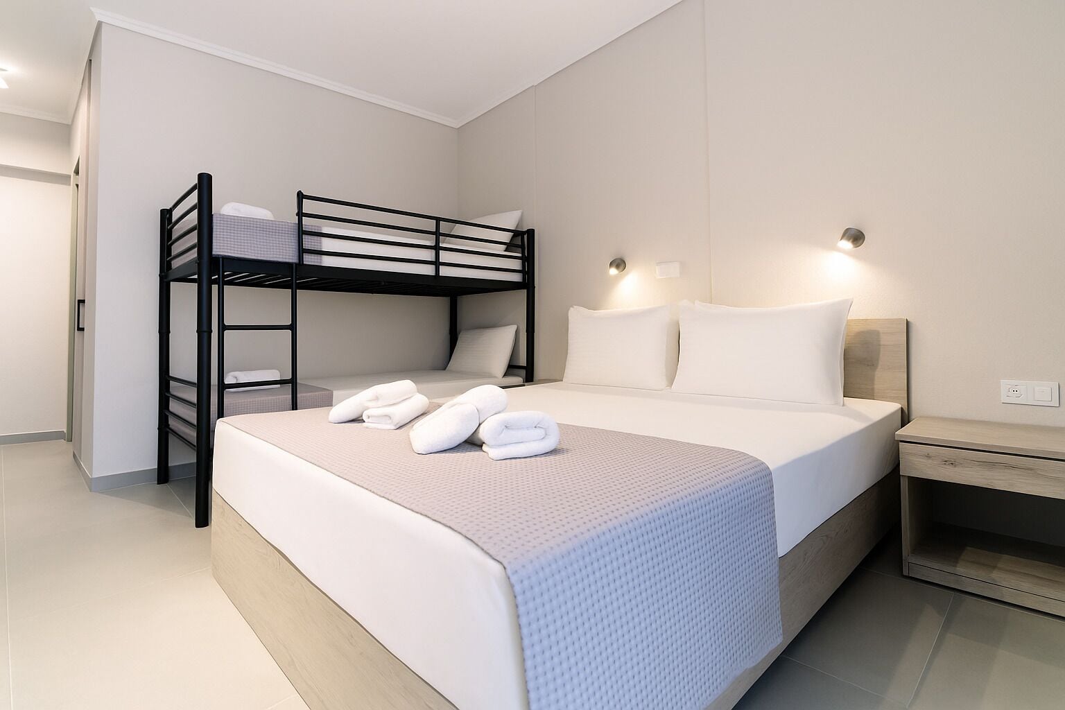 Una habitación de hotel moderna con una cama nido, una cama grande con ropa blanca, una mesita de noche y una mesilla.