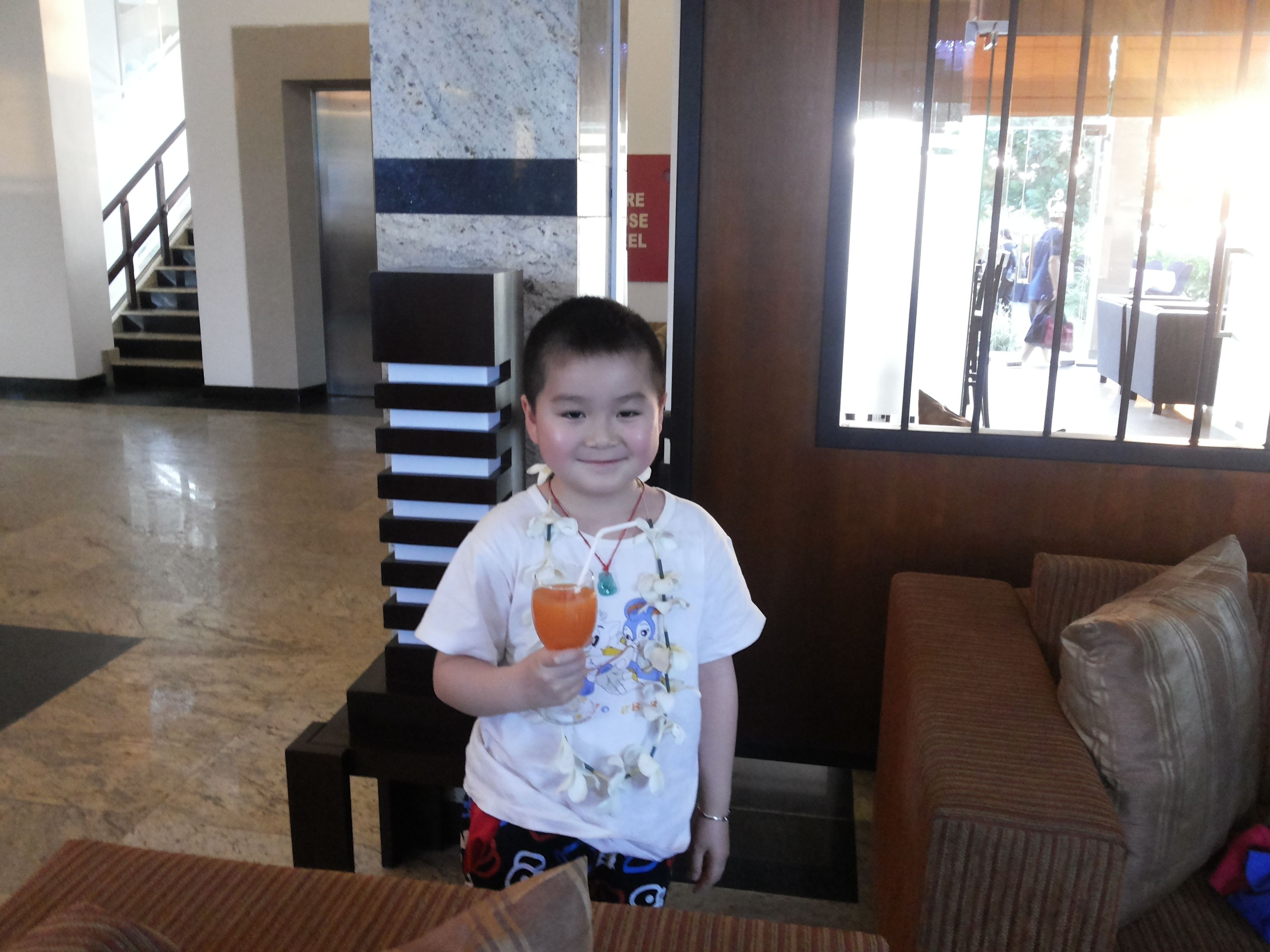 Niño sosteniendo una bebida en el vestíbulo de un hotel, con una escalera, un muro de madera y vistas al exterior.