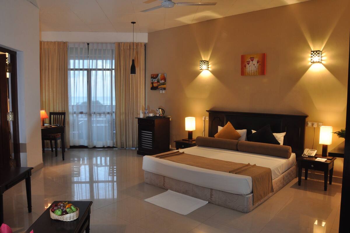 Hotelkamer met een groot bed, twee bedlampjes, een bureau met lamp, een stoel, een eettafel met een fruitmand, en uitzicht op het landschap door het raam.