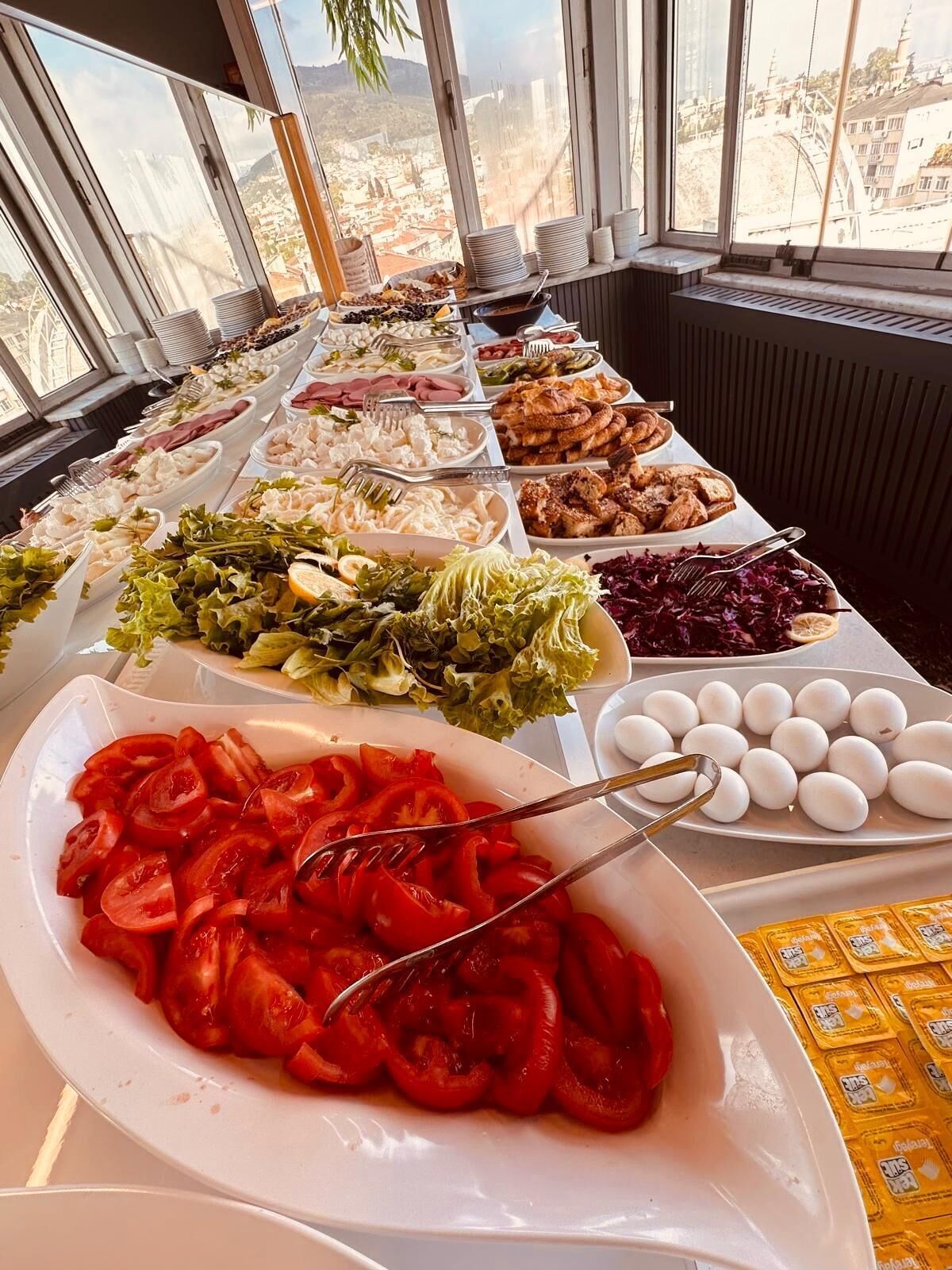 A buffet table with various food items and a view of a cityscape through large windows.