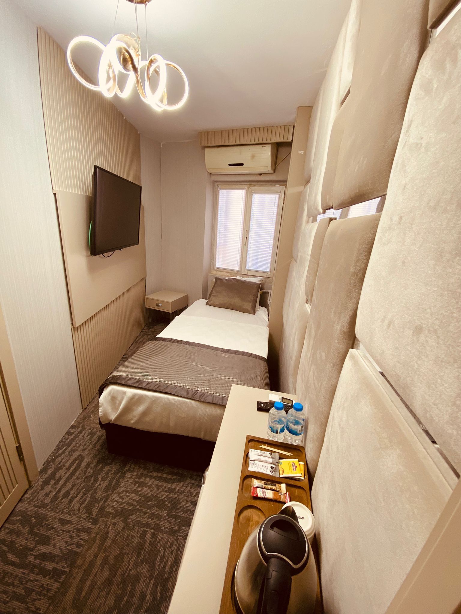 A compact hotel room with a bed, a bedside table with a kettle, a remote control, and a tray with snacks.
