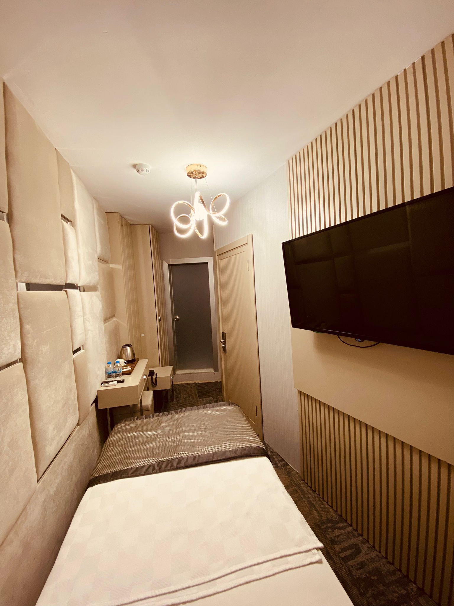 Et aflangt hotelværelse med en seng, et lille skrivebord, et fjernsyn og en usædvanlig loftlampe.