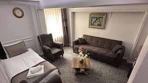 Una habitación de hotel con una cama, dos sillones, un sofá, una mesa de centro con flores, un cuadro en la pared y un reloj.