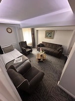 Una habitación de hotel con una cama, dos sillones, un sofá, una mesa de centro con flores, un cuadro en la pared y un reloj.