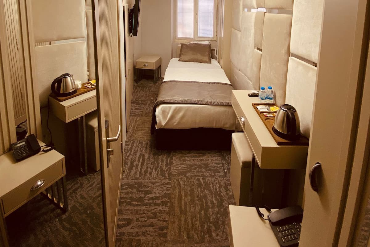 A compact hotel room with a bed, bedside table, and a desk with a chair. There is a window with blinds, a ceiling light fixture, and a wall-mounted TV.