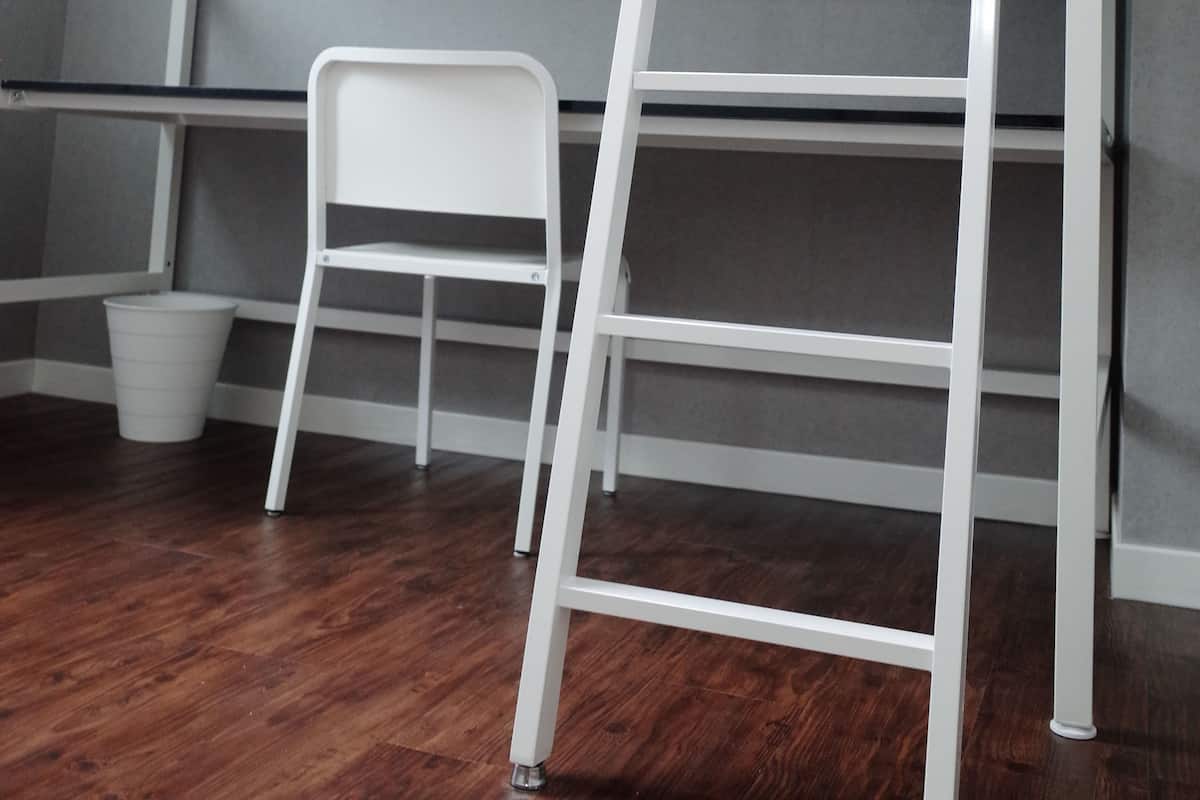 A bunk bed with a ladder, a white chair, and a white trash bin in a room with wooden flooring.