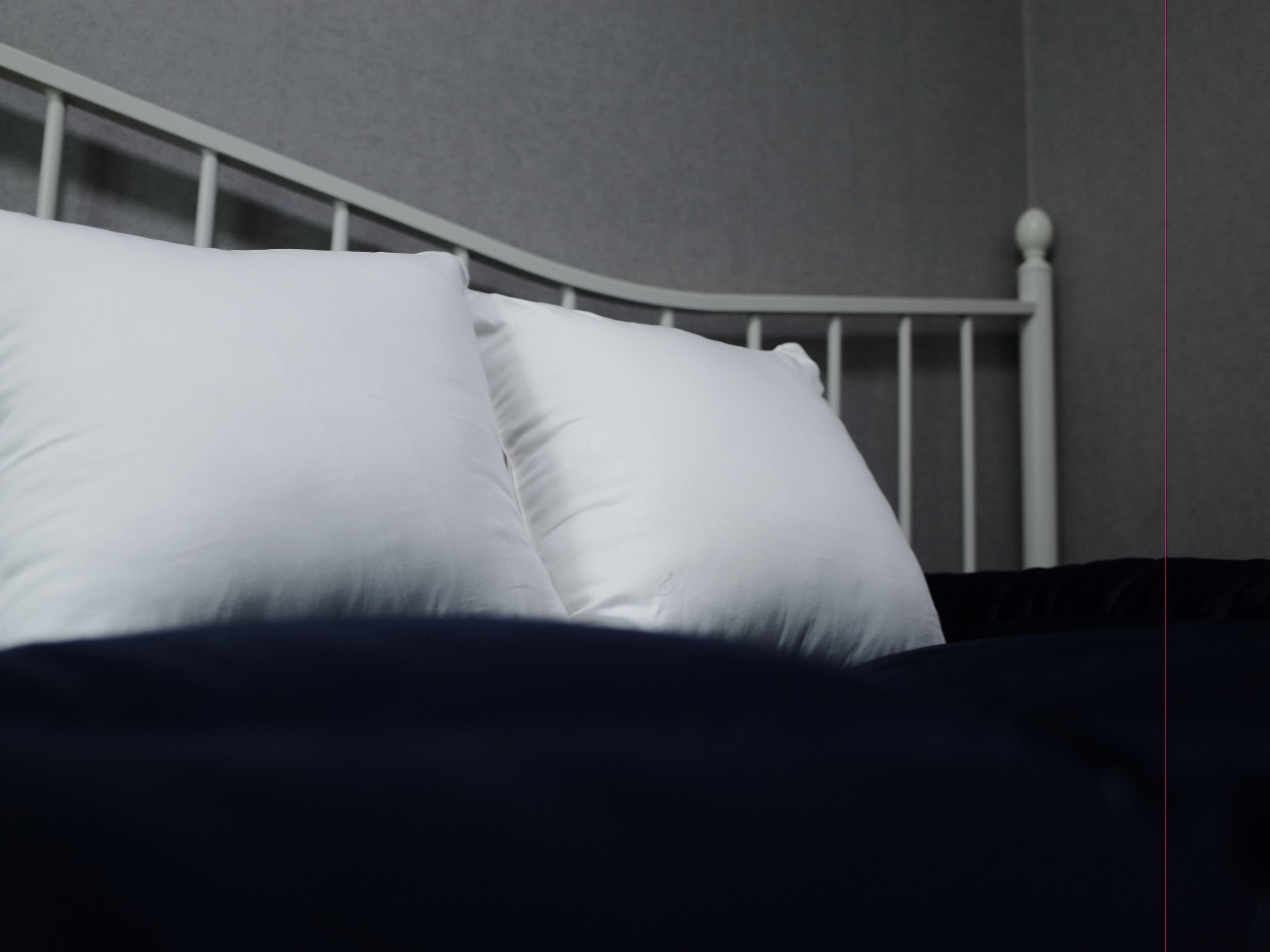 A bed with white pillows and a dark-colored duvet, a white metal headboard, and a plain wall in the background.