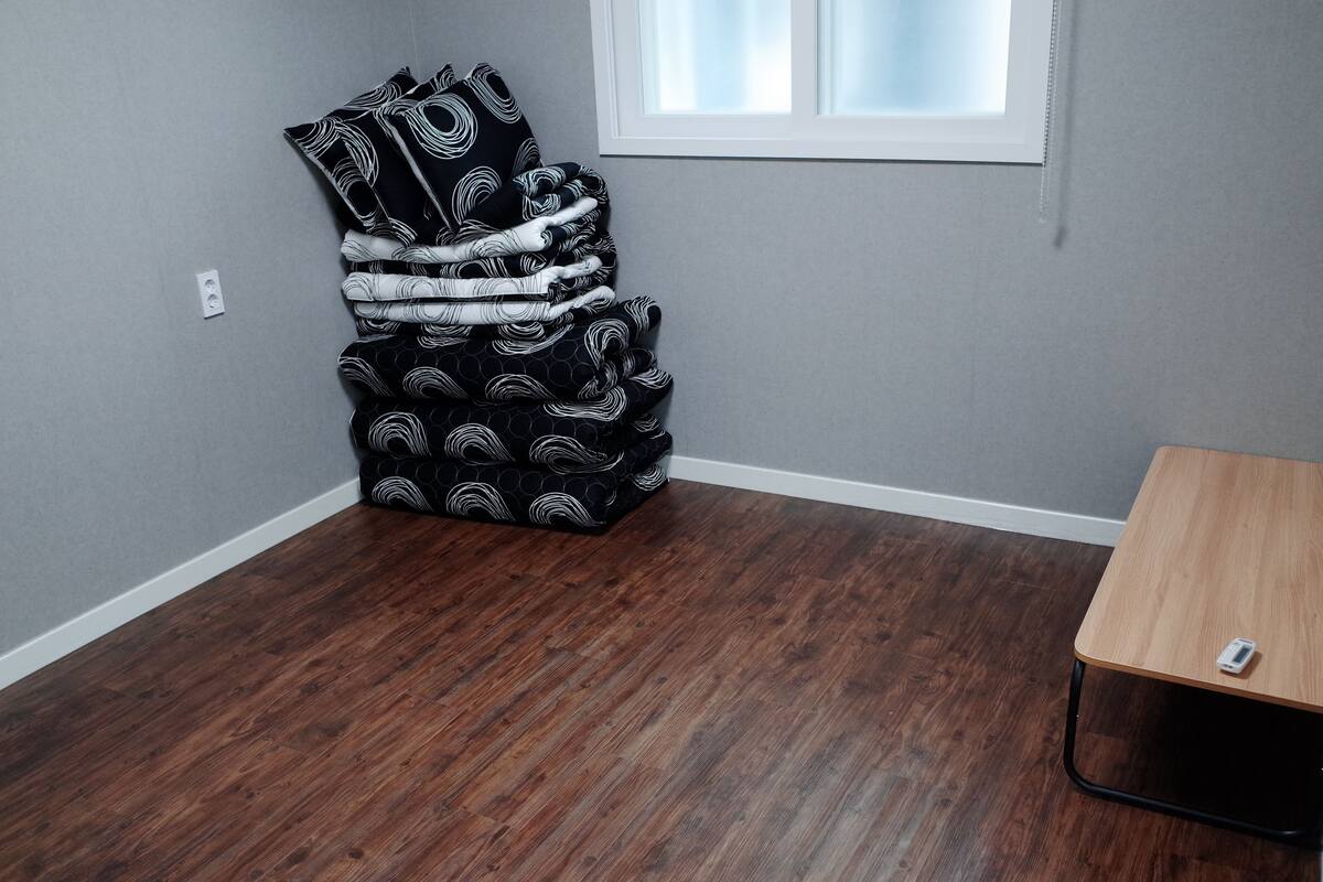 A room with a dark wood floor, a corner with a stack of patterned cushions, a small table, and a window with blinds.