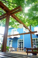 Una estructura de pérgola de madera con farolillos de papel colgantes, un edificio azul con ventanas amplias y plantas en macetas.