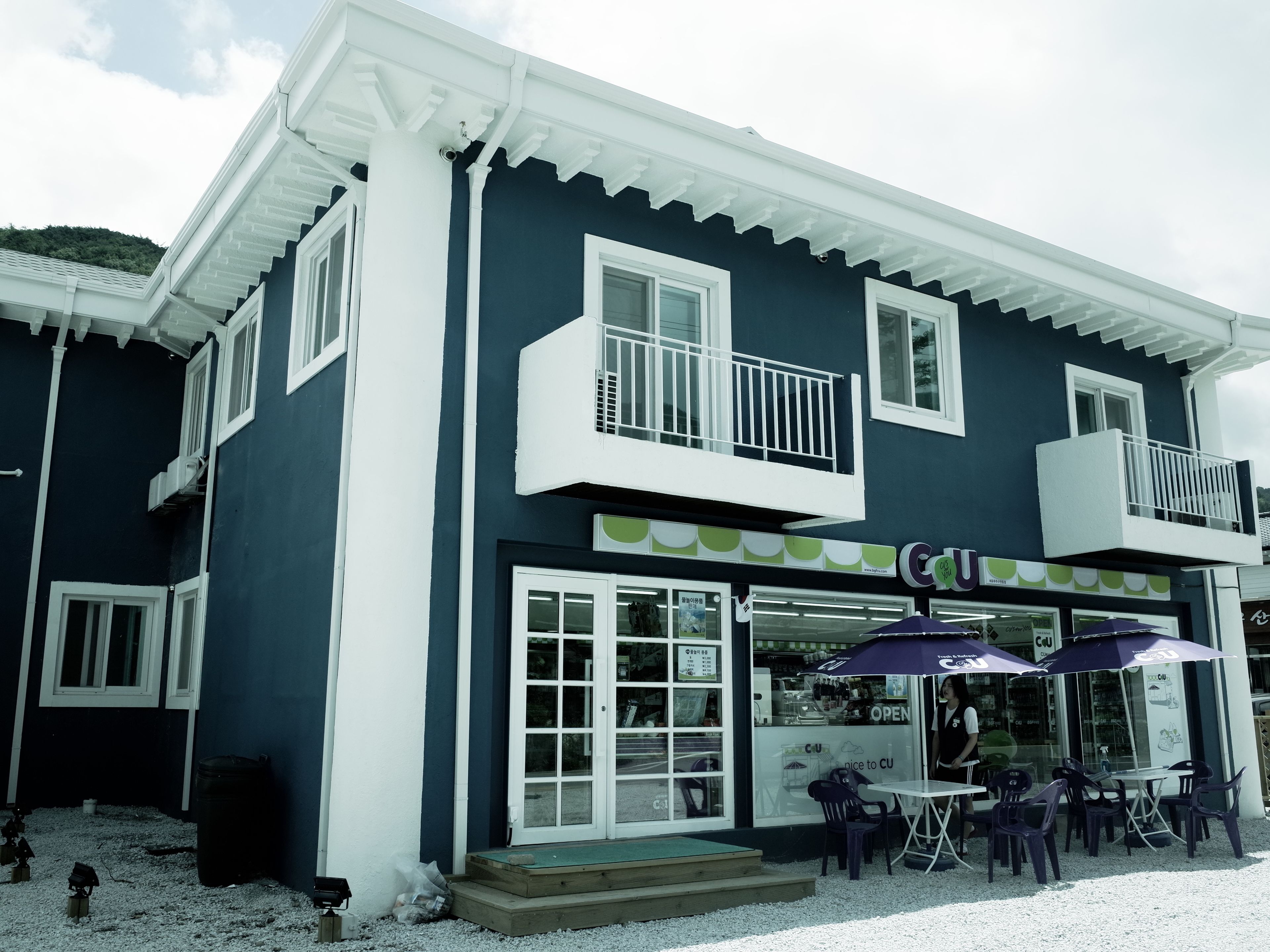 A two-story building with a dark blue exterior and white trim. It features a storefront with large windows and a sign that reads "CU". Outside, there are tables and chairs arranged for outdoor seating.