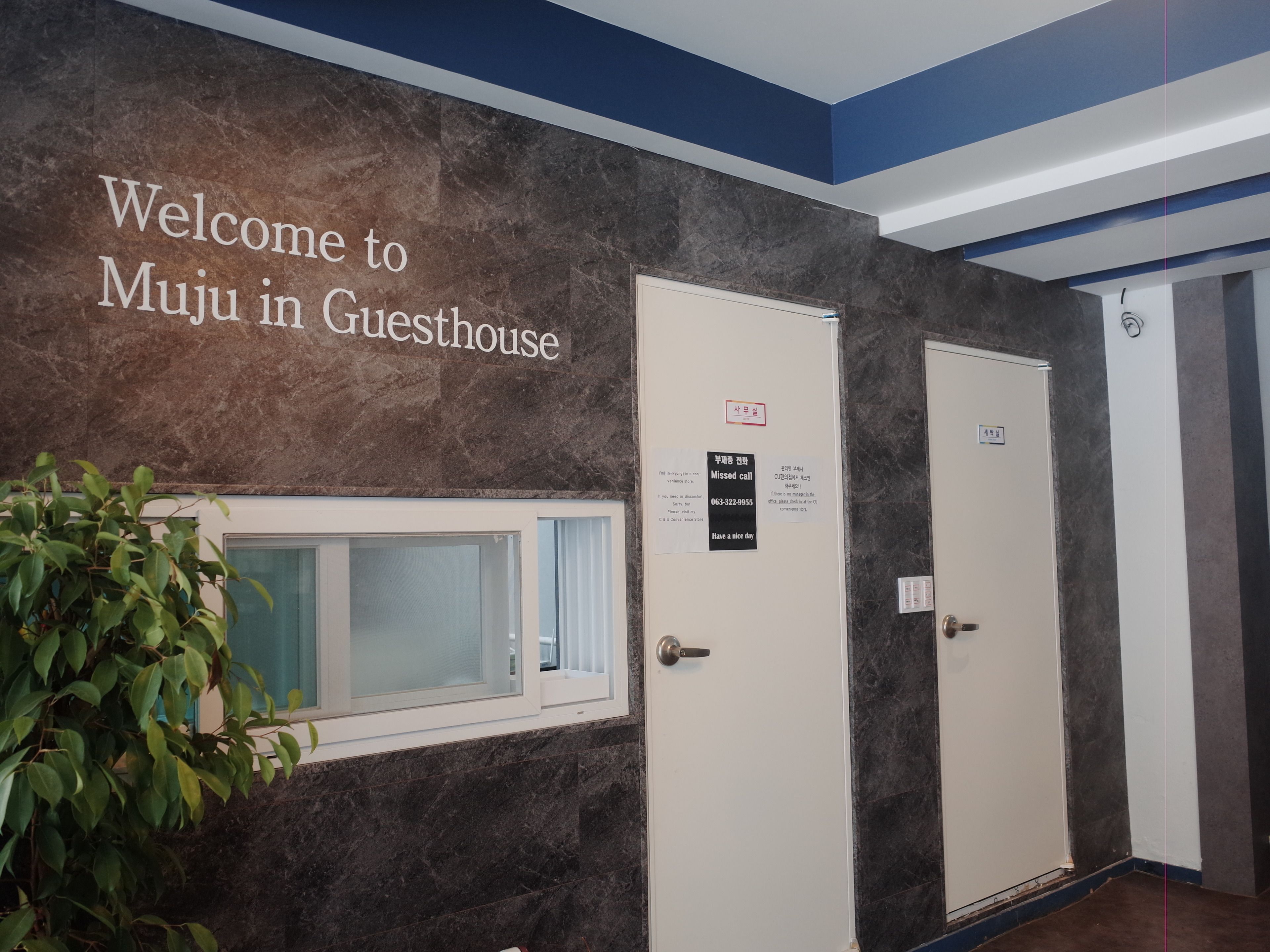 Uma área de recepção com um letreiro 'Bem-vindo ao Muju in Guesthouse', um balcão de atendimento e duas portas com a indicação 'Ligação não atendida'.