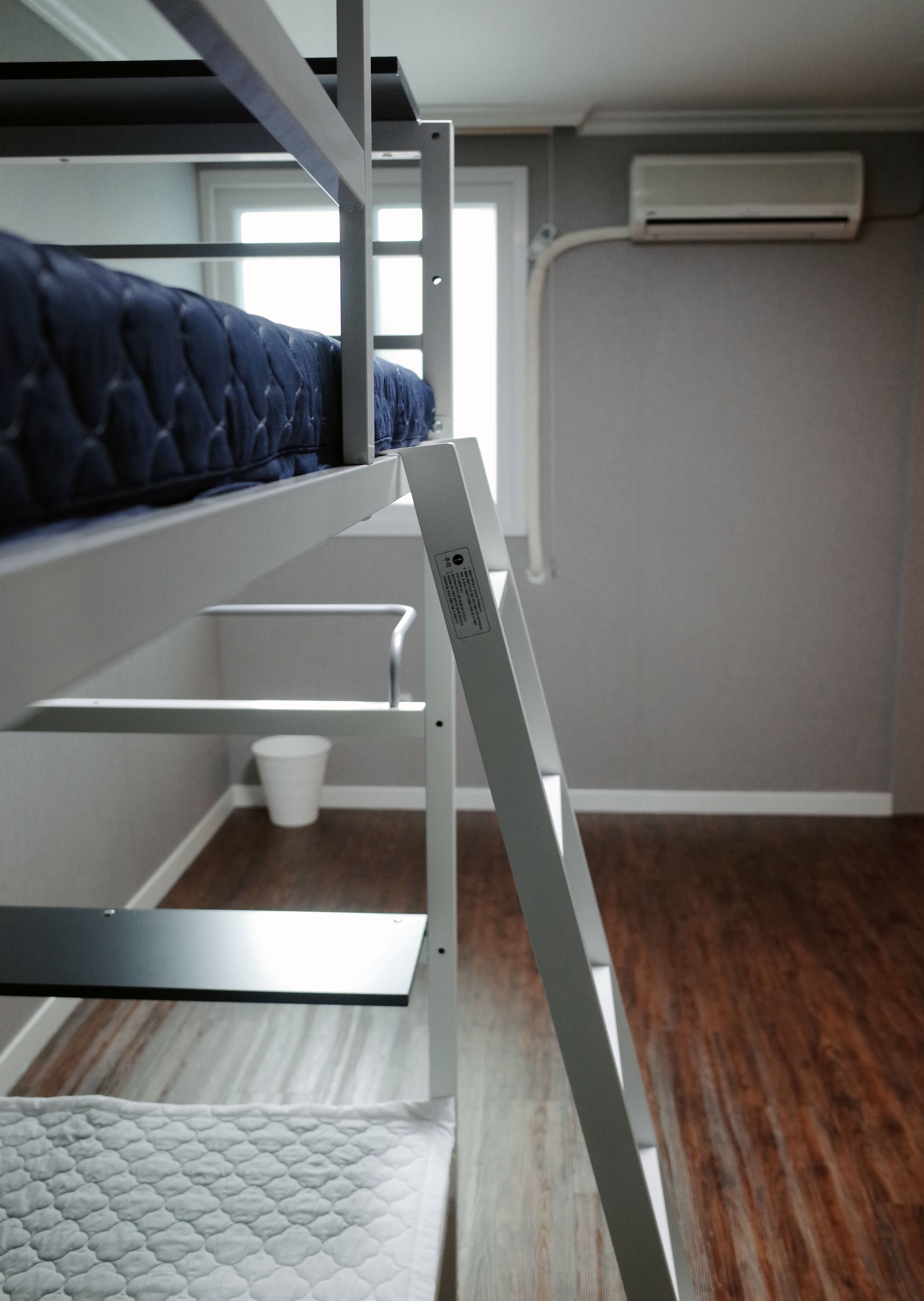 A bunk bed with a white metal frame and a quilted blue mattress. There is a white bucket on the floor and a white mattress on the lower bunk.