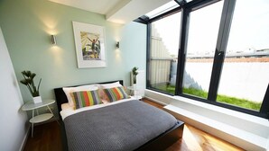 Um quarto moderno com uma cama grande, uma mesinha de cabeceira, uma planta em vaso e uma janela com vista para o exterior.