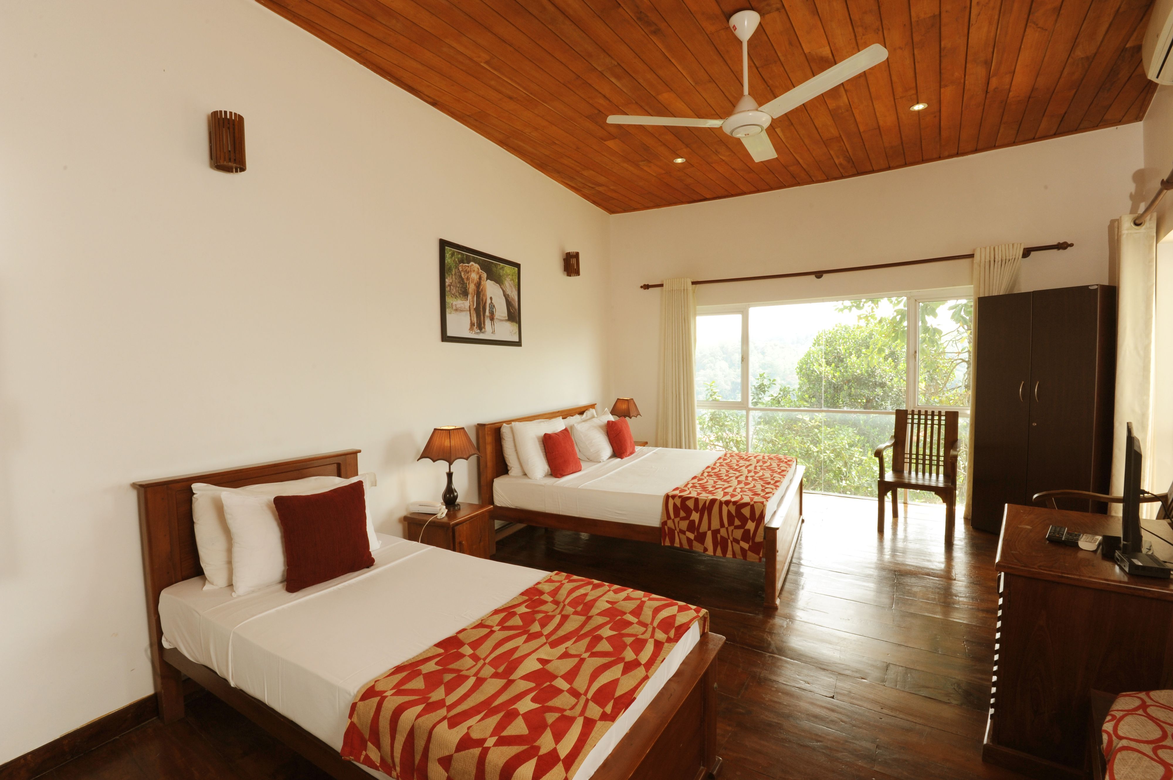 A hotel room with two beds, wooden flooring, a ceiling fan, a large window with curtains, and a desk with a television.
