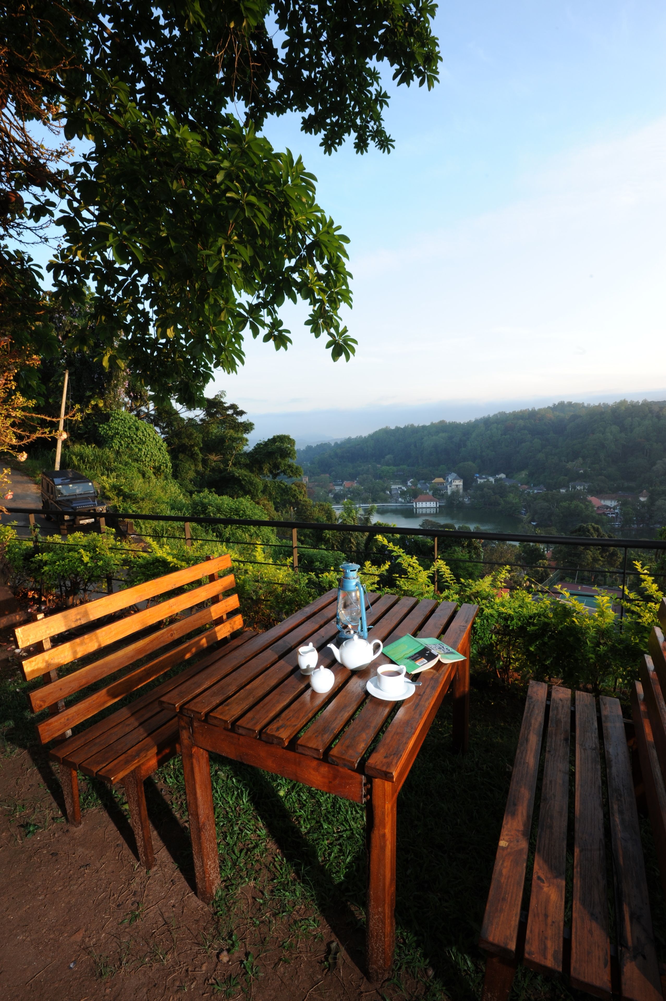 A wooden table with a white cup and saucer, a blue water bottle, and a green menu on a balcony overlooking a scenic landscape with hills and a river.