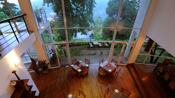A restaurant with high ceilings and large windows offering a view of a garden and trees. Tables are set with white tablecloths and red napkins. - Hotel See Kandy (Kandy)