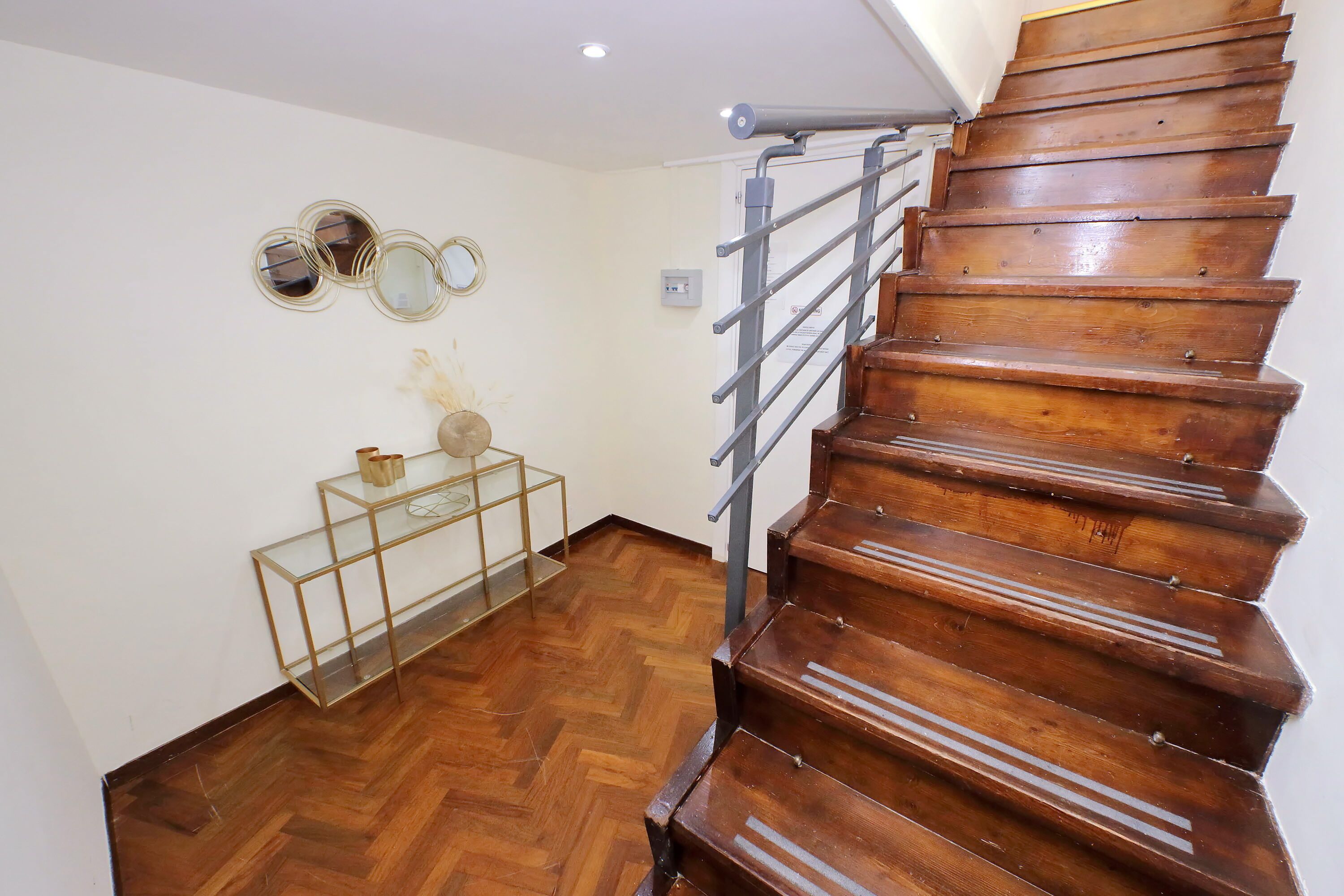 A wooden staircase with a metal handrail, a glass console table with decorative items, and a herringbone-patterned wooden floor.