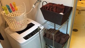 A laundry area with a washing machine, a basket of colorful clothespins, and a shelf with folded towels. - Sunlife Yamashina Apartment (Kyoto)