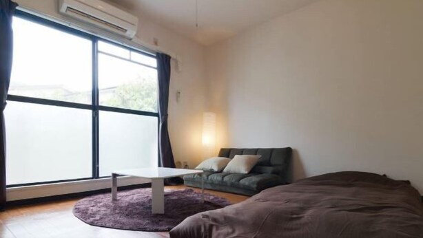 A room with a large window, a white table, a green sofa, and a brown bed. - Sunlife Yamashina Apartment (Kyoto)