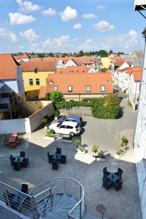 A courtyard with outdoor seating, a white car parked in the middle, and buildings with red-tiled roofs in the background.
