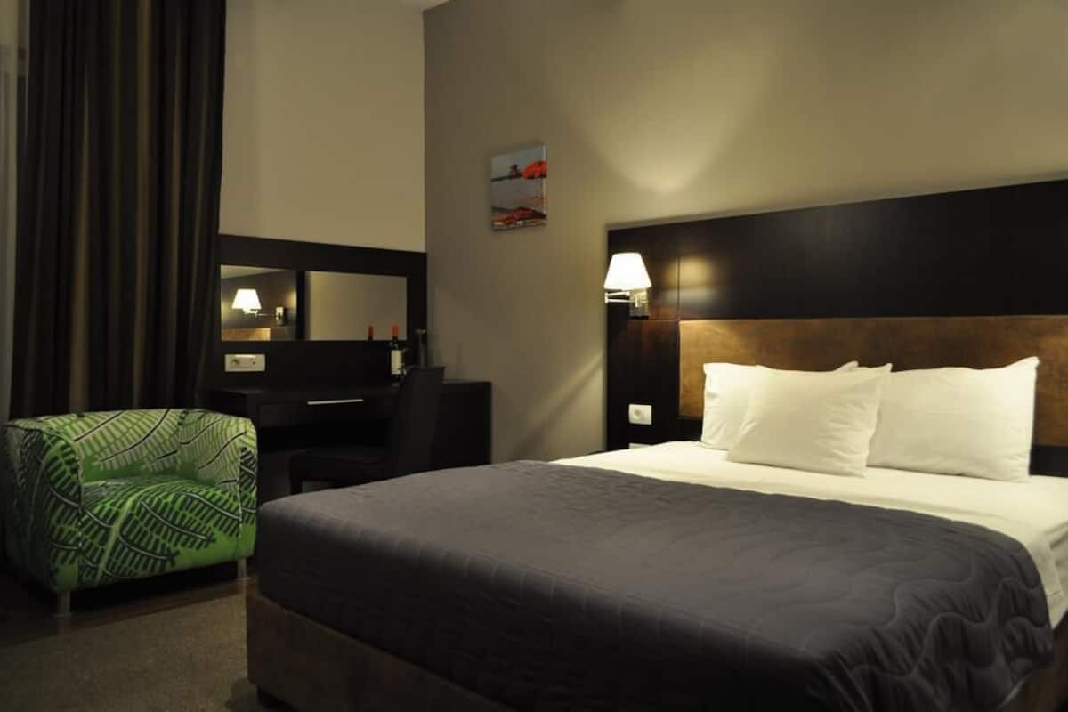 Una habitación de hotel con una cama grande, un escritorio con silla, un sillón verde estampado, una lámpara de pared y un cuadro.