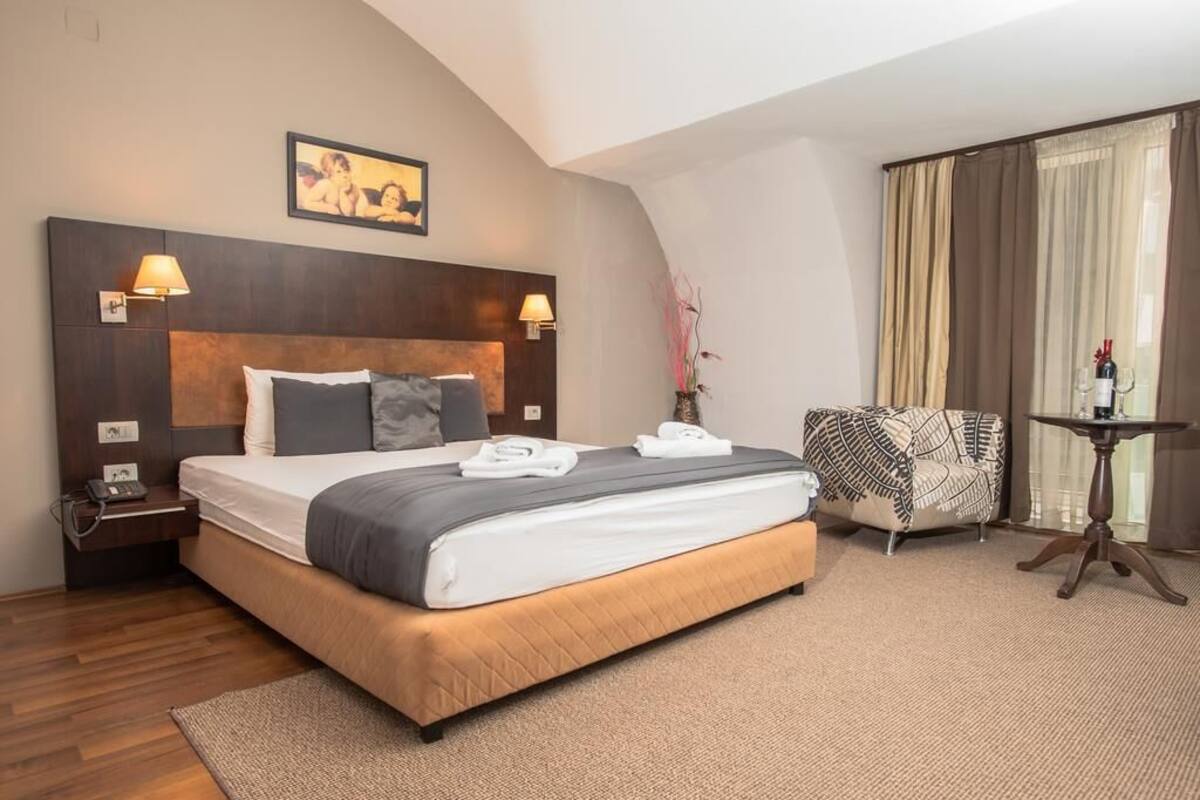 Una habitación de hotel con una cama grande, dos lámparas de noche, una mesita de noche, una silla, una pequeña mesa y una ventana con cortinas.