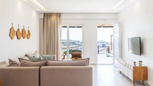 A modern living room with a sectional sofa, a flat-screen TV mounted on the wall, and a view of the city through large windows. - Evi Suites & Villas (Mykonos)