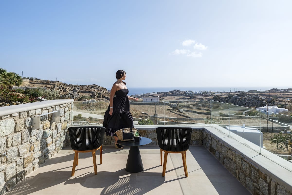 A terrace with stone walls, two black chairs, a small table, and a woman in a black dress.