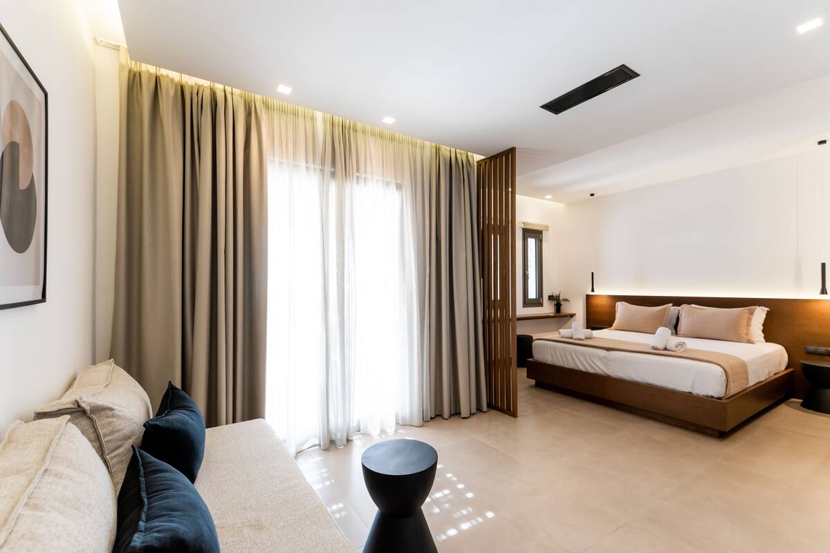 A modern hotel room with a large bed, a sofa, a small round table, and a large window with curtains.
