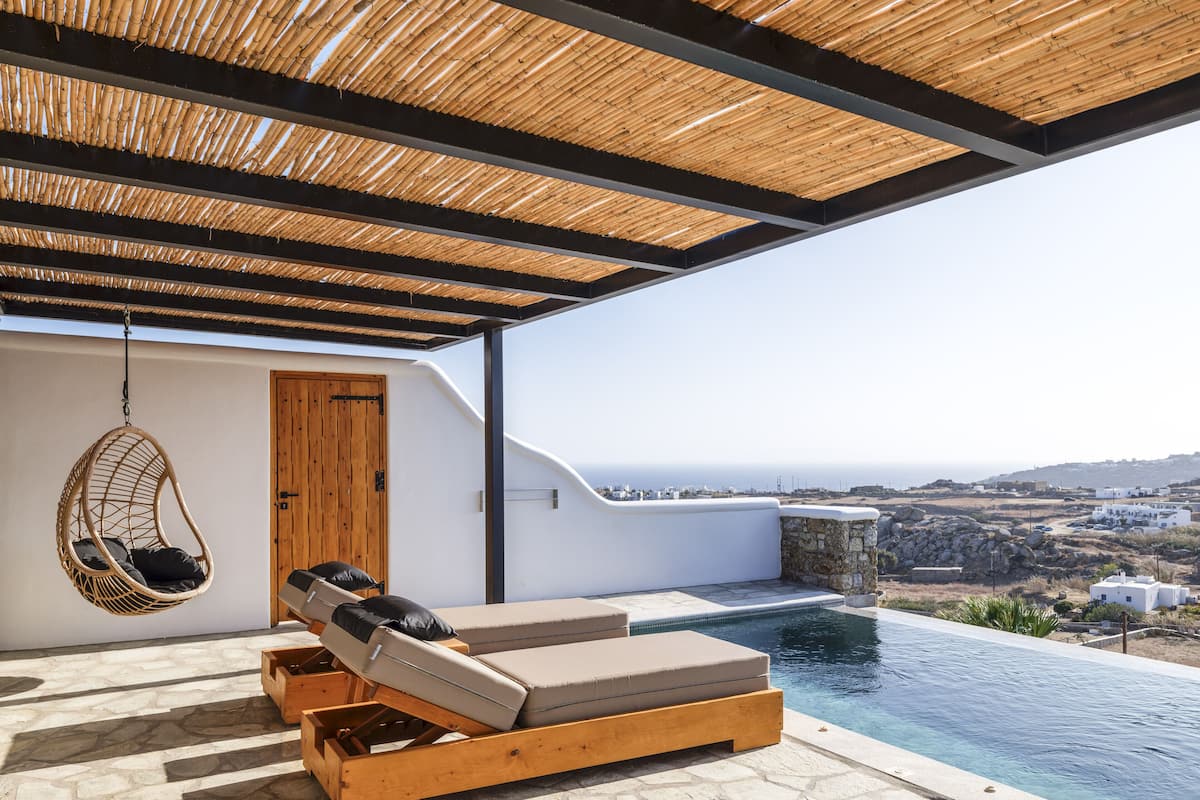 A modern outdoor lounge area with a stone floor, wooden loungers, a hanging chair, and a view of the sea.