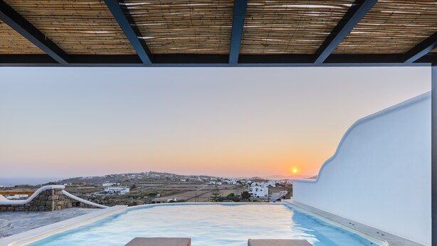 A poolside lounge with a view of a sunset over a coastal village. - Evi Suites & Villas (Mykonos)