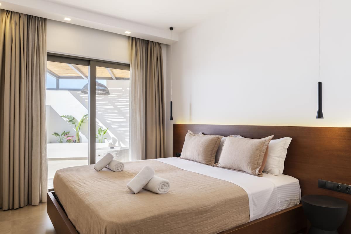 A modern hotel room with a large bed, beige curtains, a view of the outdoors, and a minimalist design.