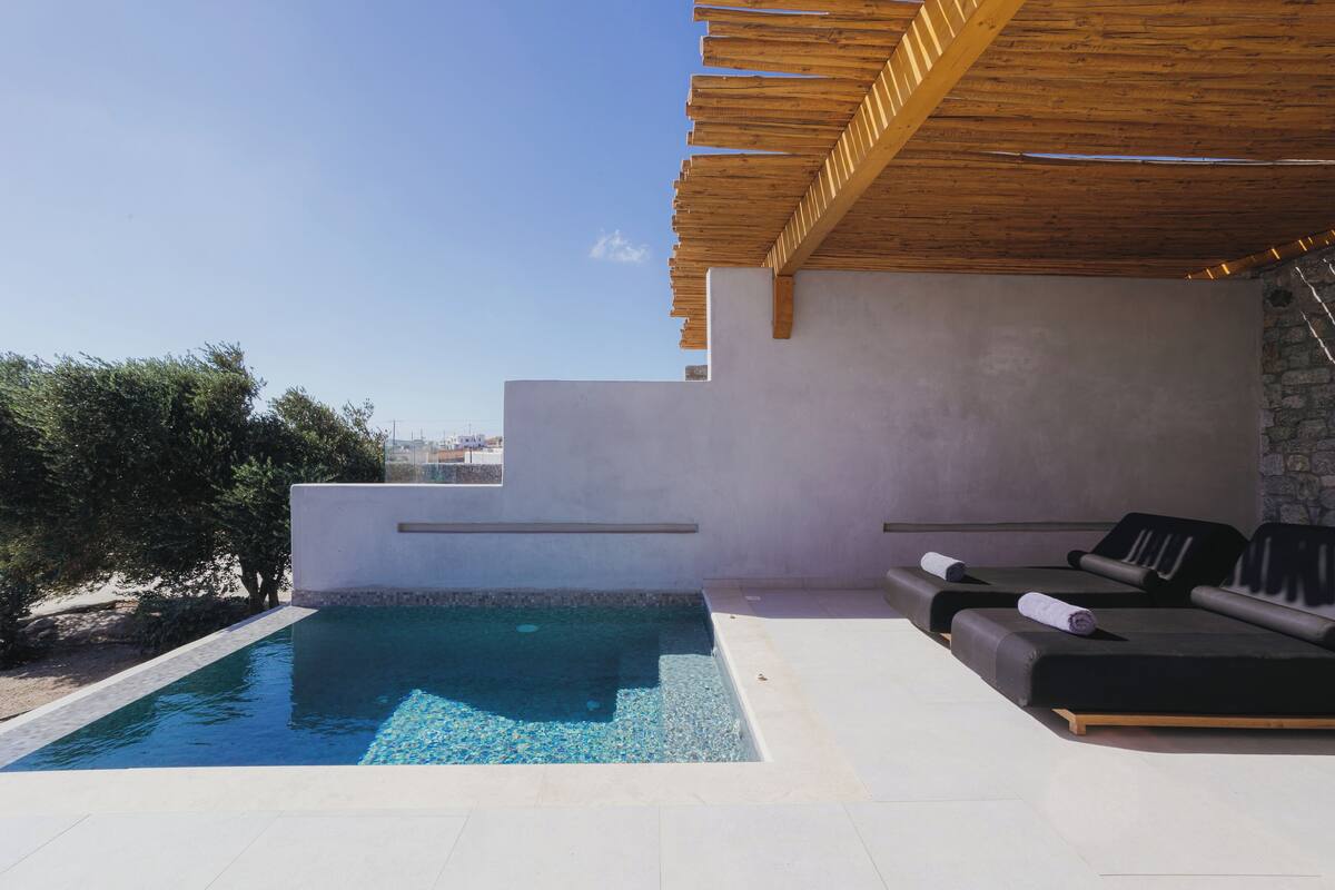 A modern outdoor pool area with a wooden pergola, a stone wall, and a seating area with cushions.