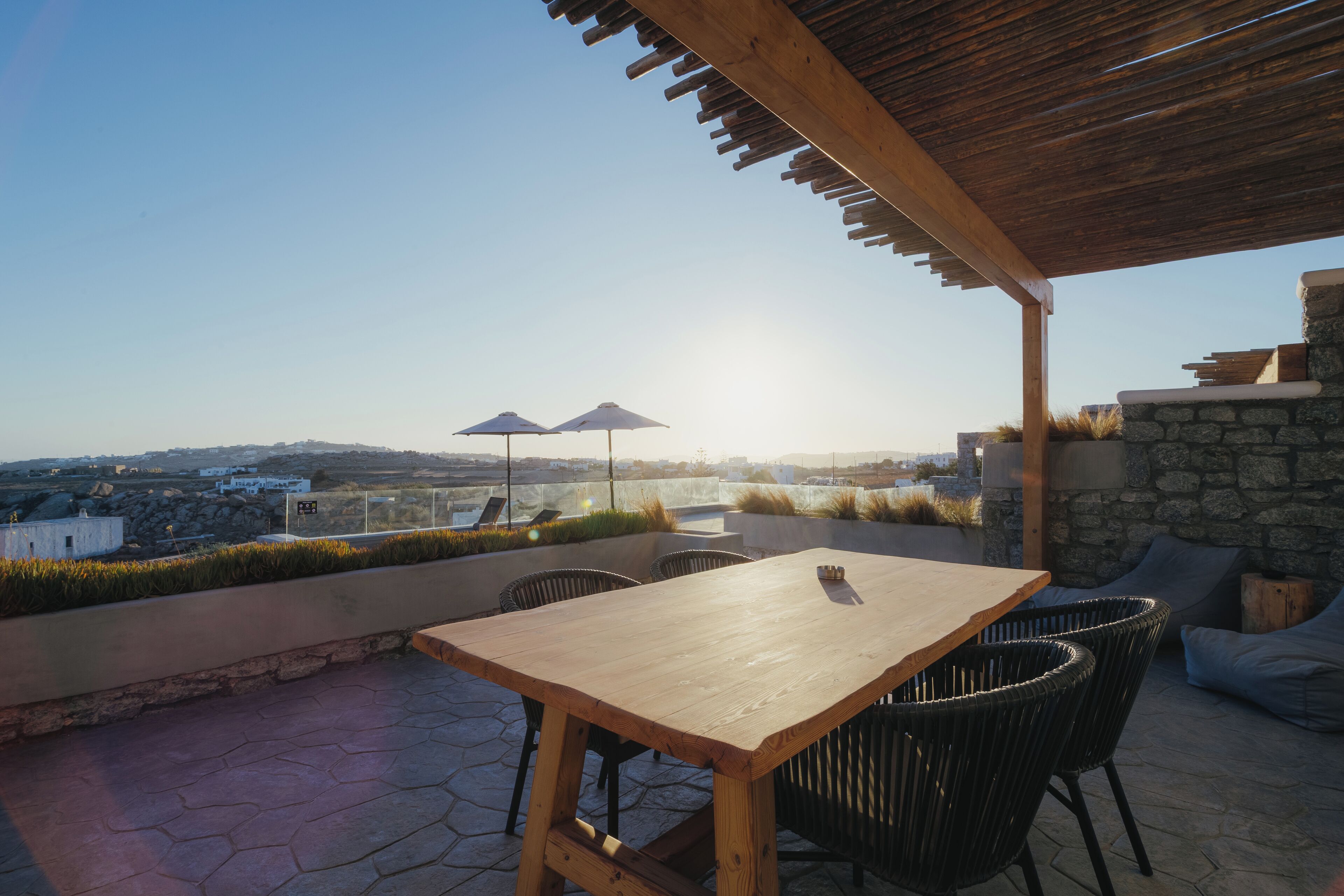 Een terras buiten met een houten tafel en stoelen, stenen muren en twee parasols.