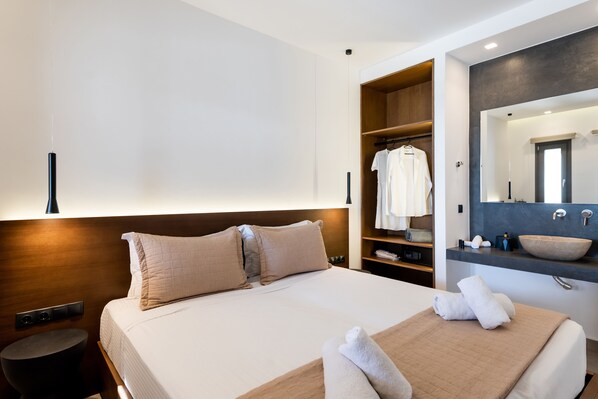 A modern hotel room with a large bed, a walk-in closet, a bathroom with a large mirror and sink, and a bedside table with a lamp. - Evi Suites & Villas (Mykonos)