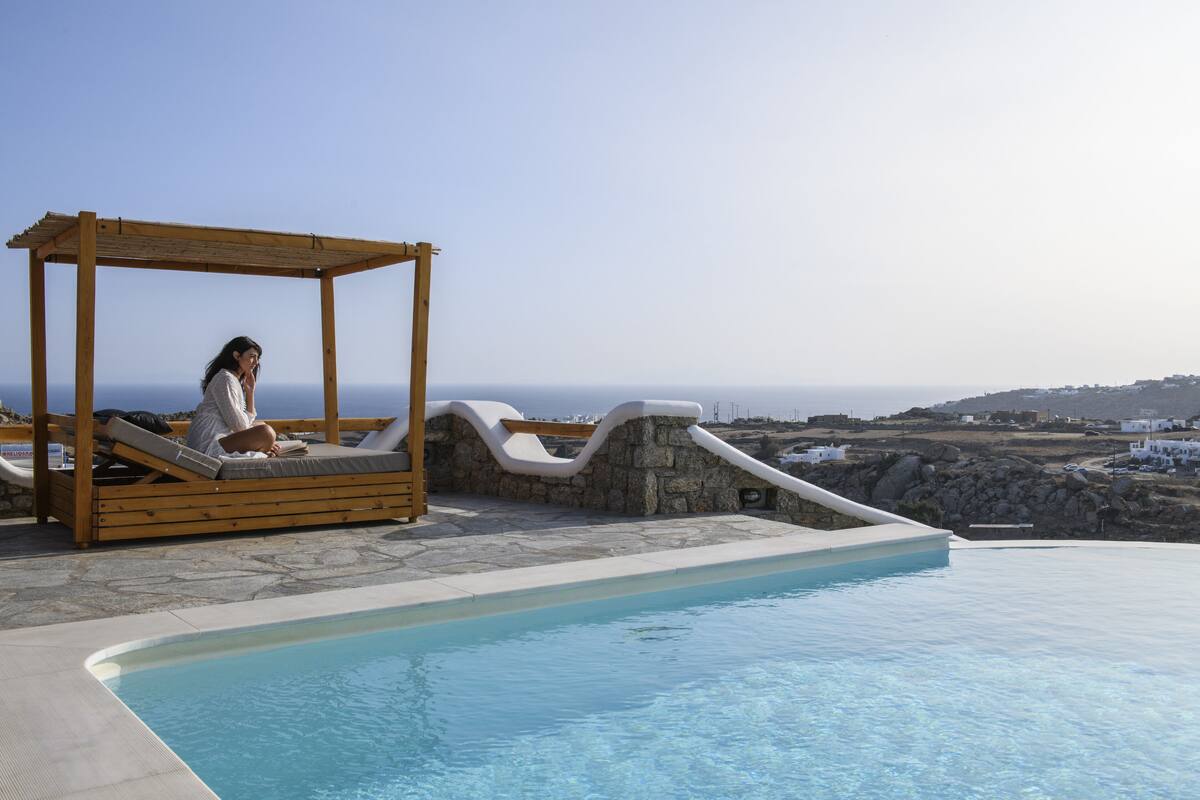 A wooden gazebo with a person relaxing by a pool with a view of the sea and distant buildings.