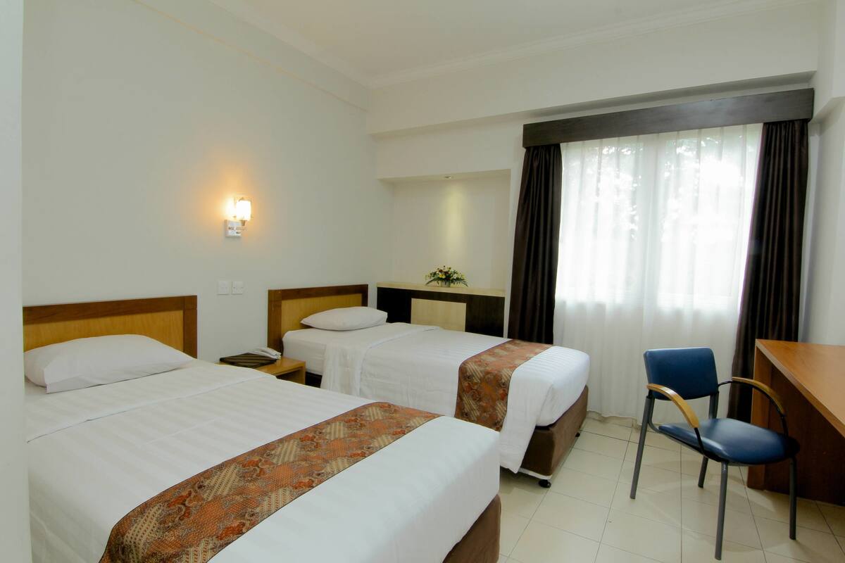 A hotel room with two beds, a desk, a chair, and a nightstand with a lamp.