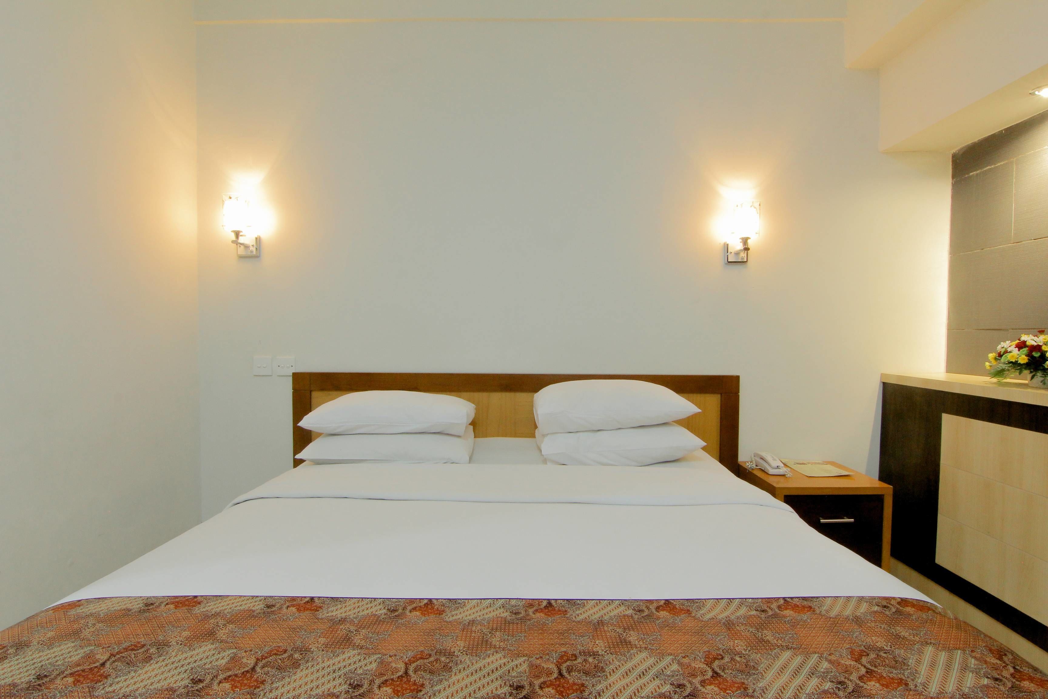 A hotel room with a neatly made bed, two bedside lamps, a nightstand with a phone, and a decorative wall panel.
