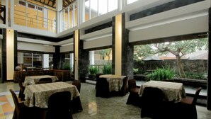 A spacious indoor area with multiple tables and chairs, a high ceiling, and large windows offering views of the outdoors. - University Hotel Yogyakarta (Yogyakarta)