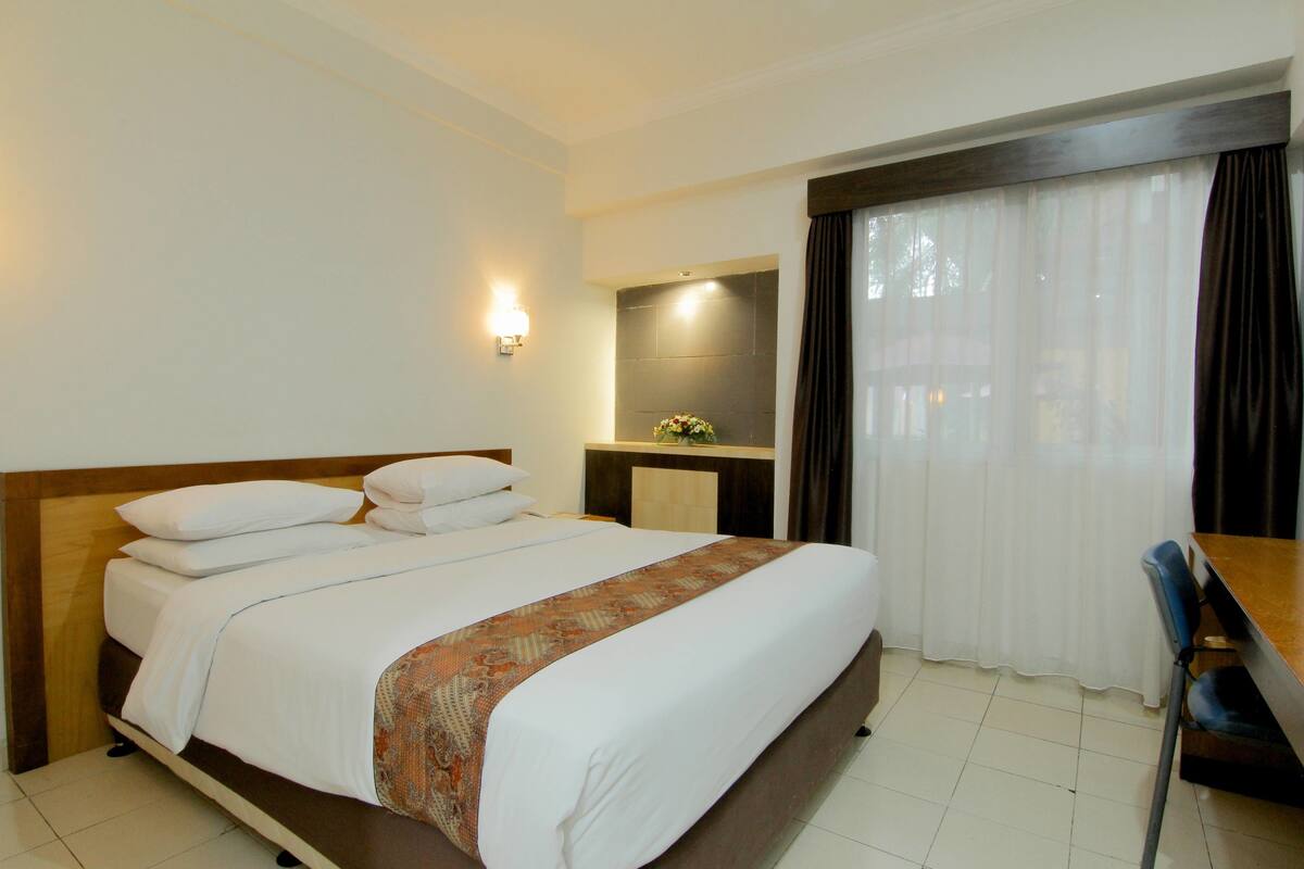 A hotel room with a large bed, a desk, and a chair. There is a window with curtains and a wall-mounted light.
