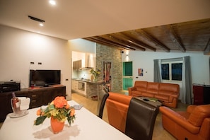 A living room with a television, a dining table with flowers, and a kitchen in the background.