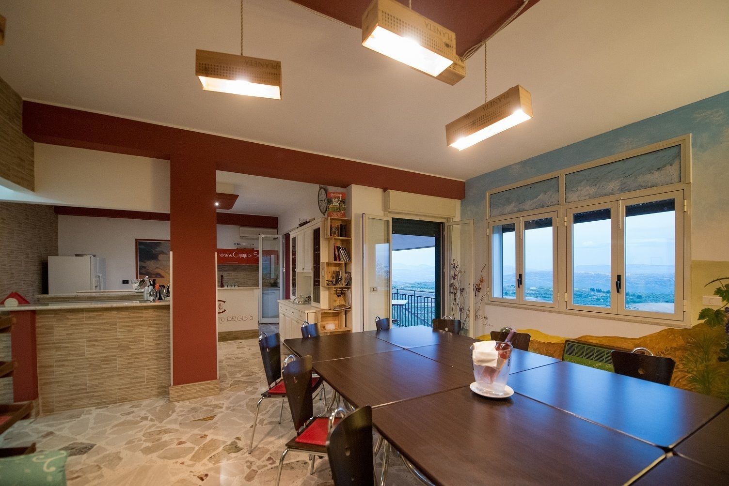 A spacious dining area with a long table and chairs, a kitchen in the background, and a view through large windows.