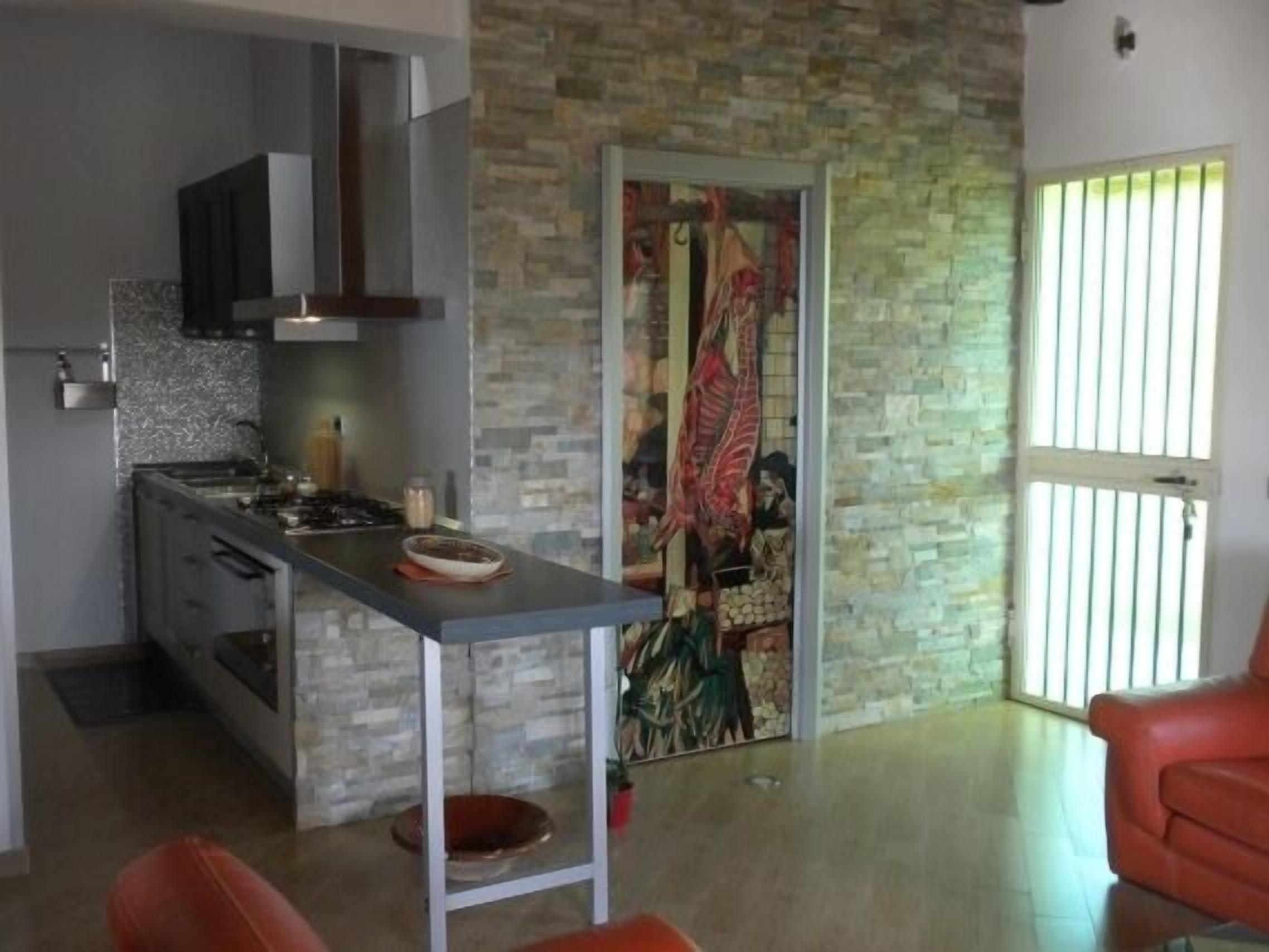 A modern kitchen with a stone wall, a dining area with a red chair, and a large window with vertical blinds.