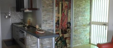 A modern kitchen with a stone wall, a dining area with a red chair, and a large window with vertical blinds.