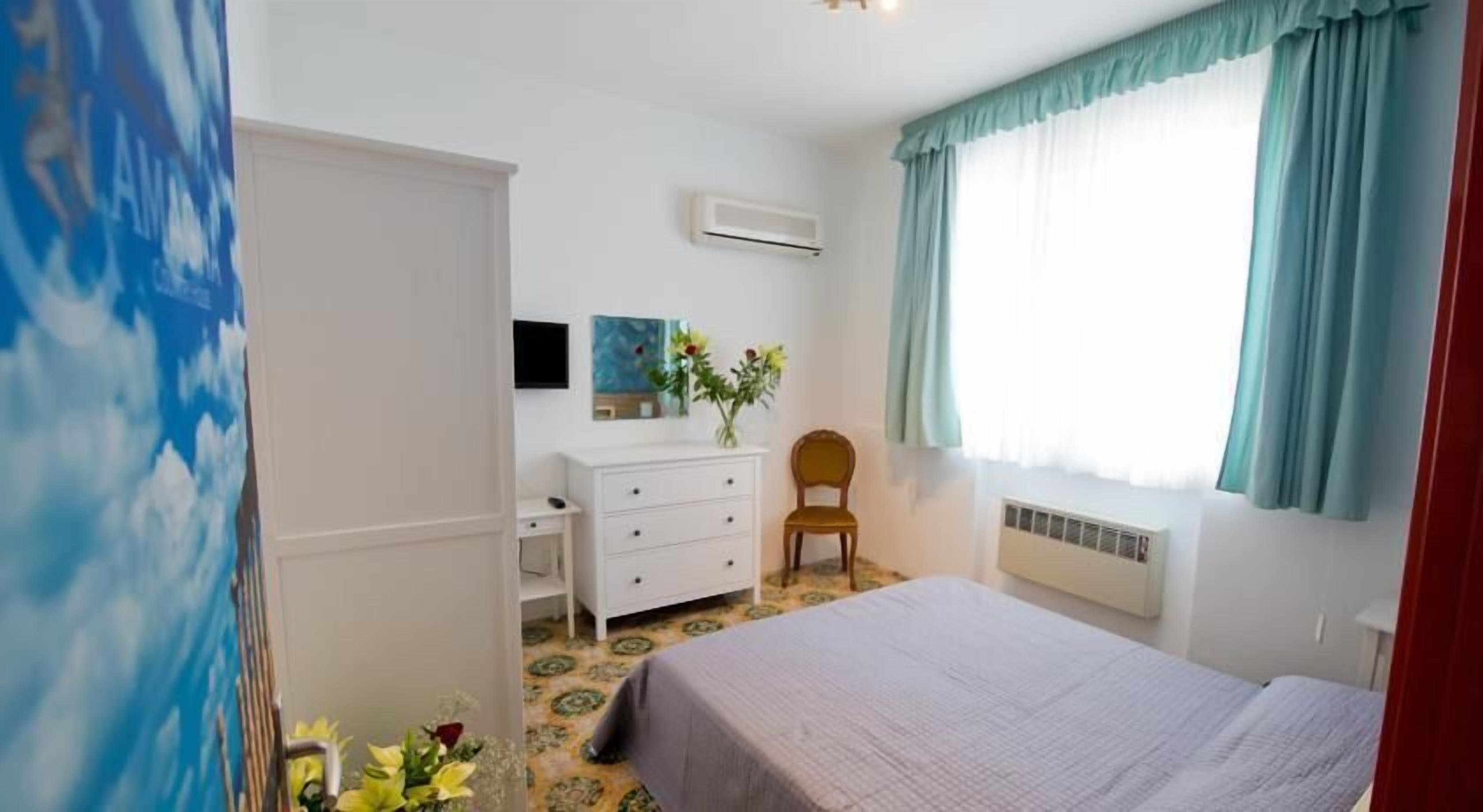 A bedroom with a bed, a dresser, a chair, an air conditioning unit, and a window with curtains.