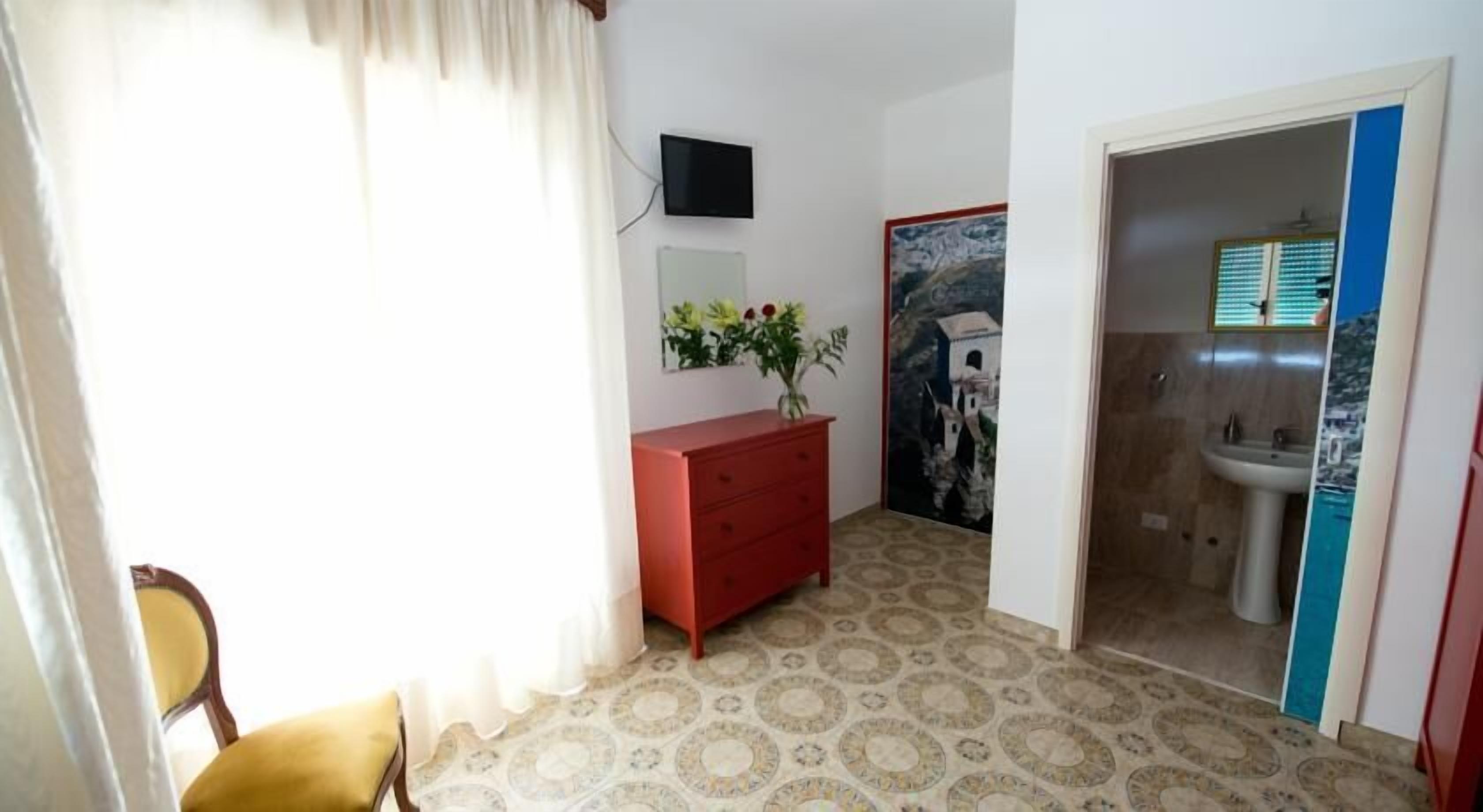 A room with a patterned floor, a red dresser, a wall-mounted TV, and a bathroom with a sink and a poster.