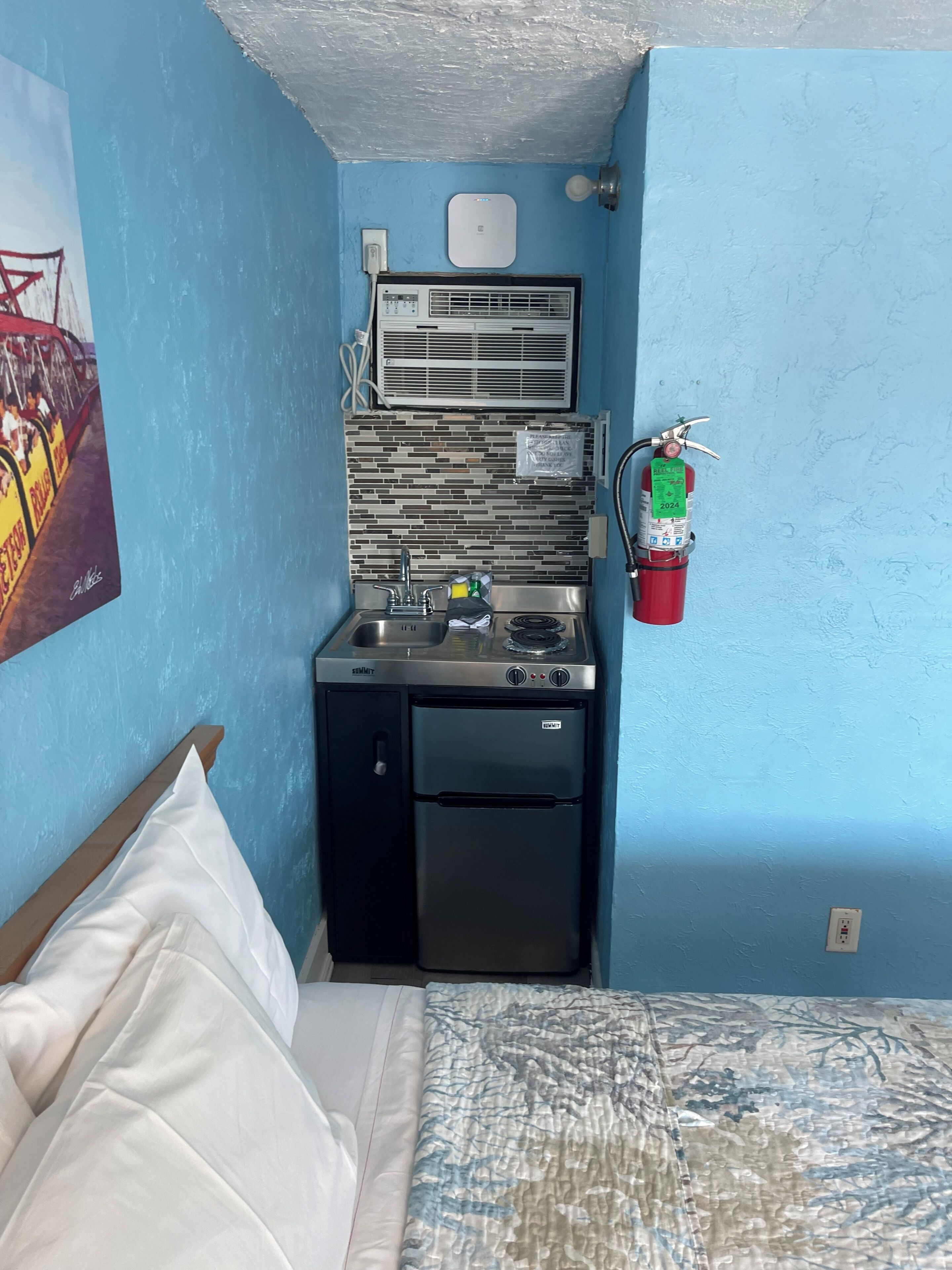 A compact room with a bed, a kitchenette with a stove, a small refrigerator, a wall-mounted air conditioner, a fire extinguisher, and a wall-mounted outlet.